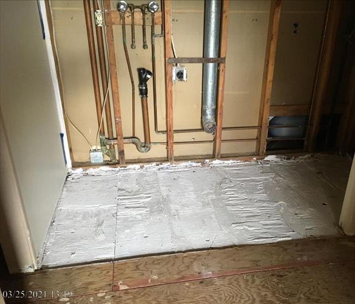 A demolished laundry room wall with removed tiles and visible laundry hookups and plumbing